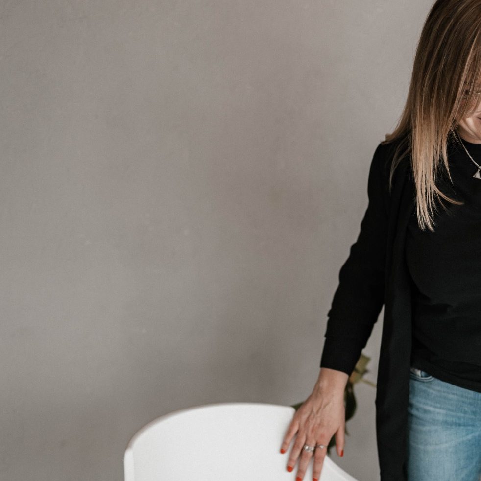 Sabine Welsch mit längeren Haaren in einem schwarzen Oberteil steht neben einem weißen Stuhl.